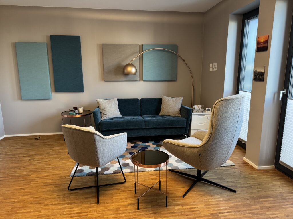 Auf dem Bild ist der Praxisraum für Paarberatung/ Paartherapie zu sehen mit einer türkisen Couch und zwei beigen Sesseln und zwei kleinen Tischen und einfarbigen Bildern an der Wand.